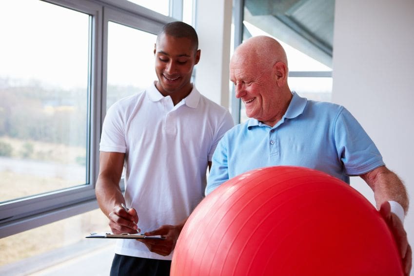 z-1QtSvU Rehab specialist looking at clipboard together with a client that is holding a large red fitness ball